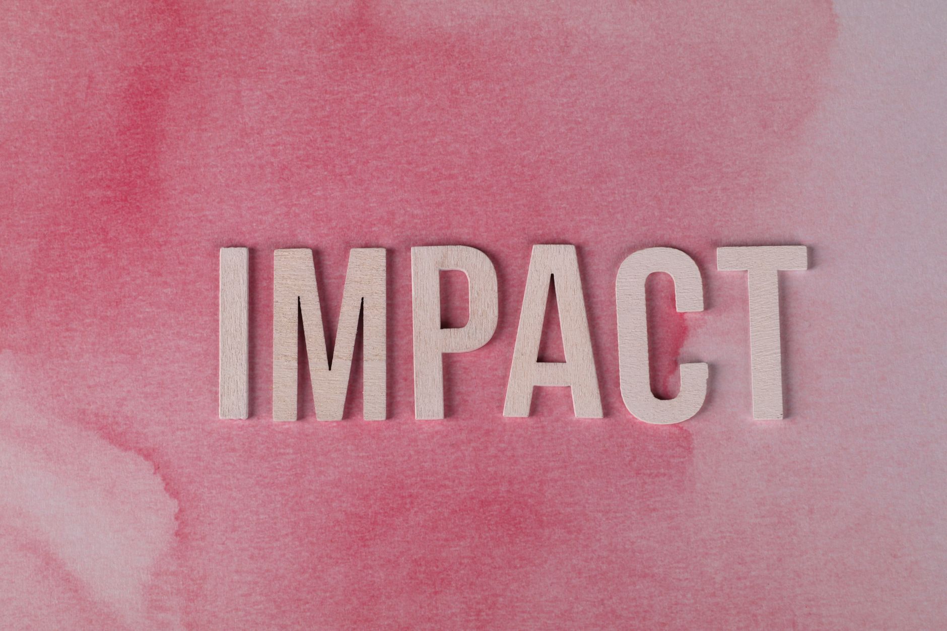 Wooden letters spelling 'IMPACT' against a textured pink watercolor backdrop, symbolizing influence and change.