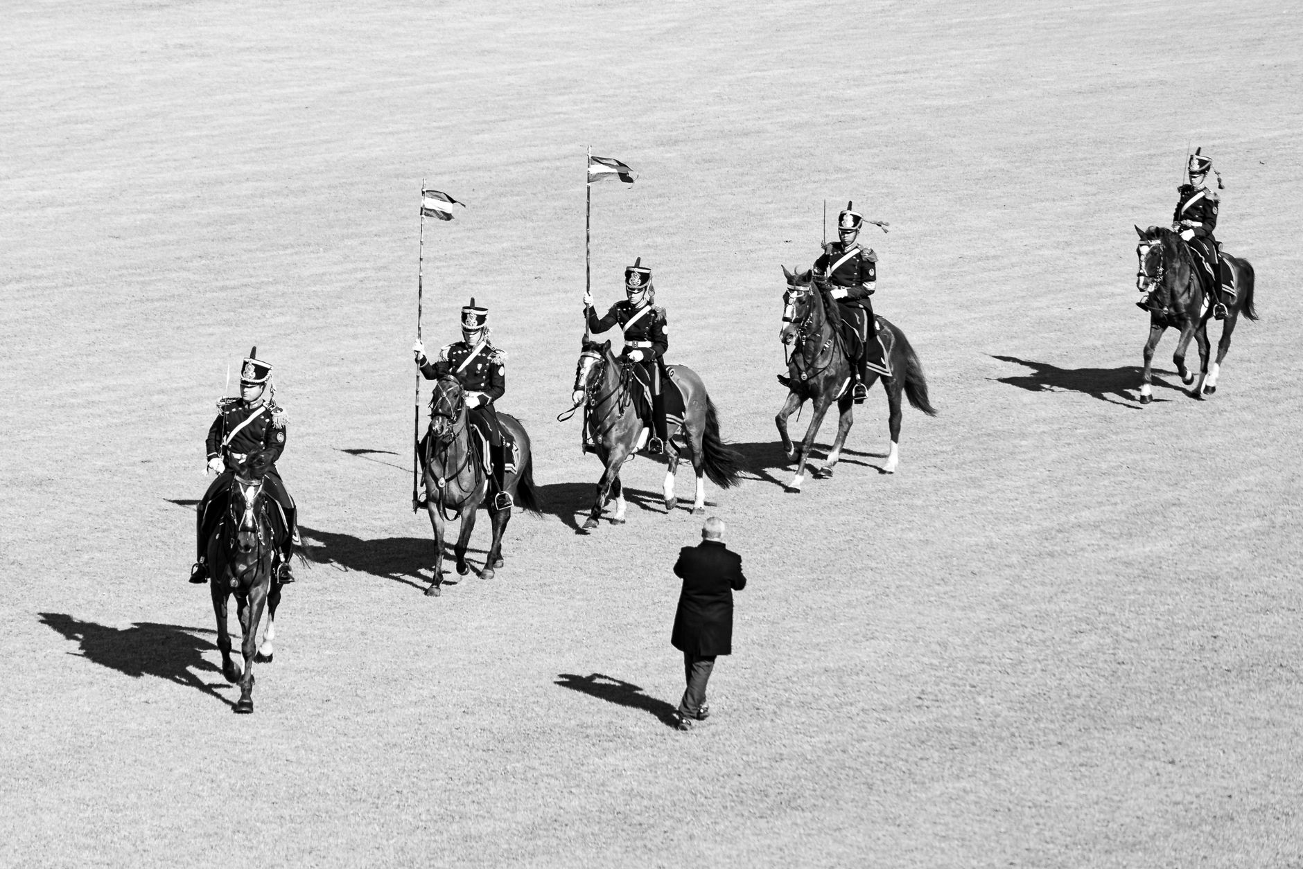 Monochrome image of uniformed riders on horses in formation during a parade.