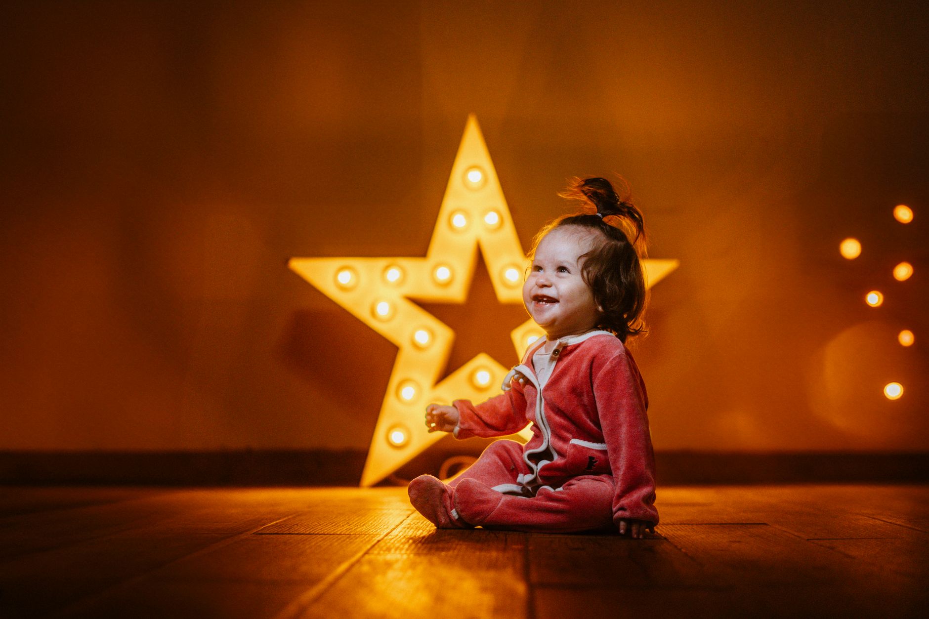 A joyful baby in red pajamas sits with illuminated star lights in the background, creating a warm and cheerful atmosphere.