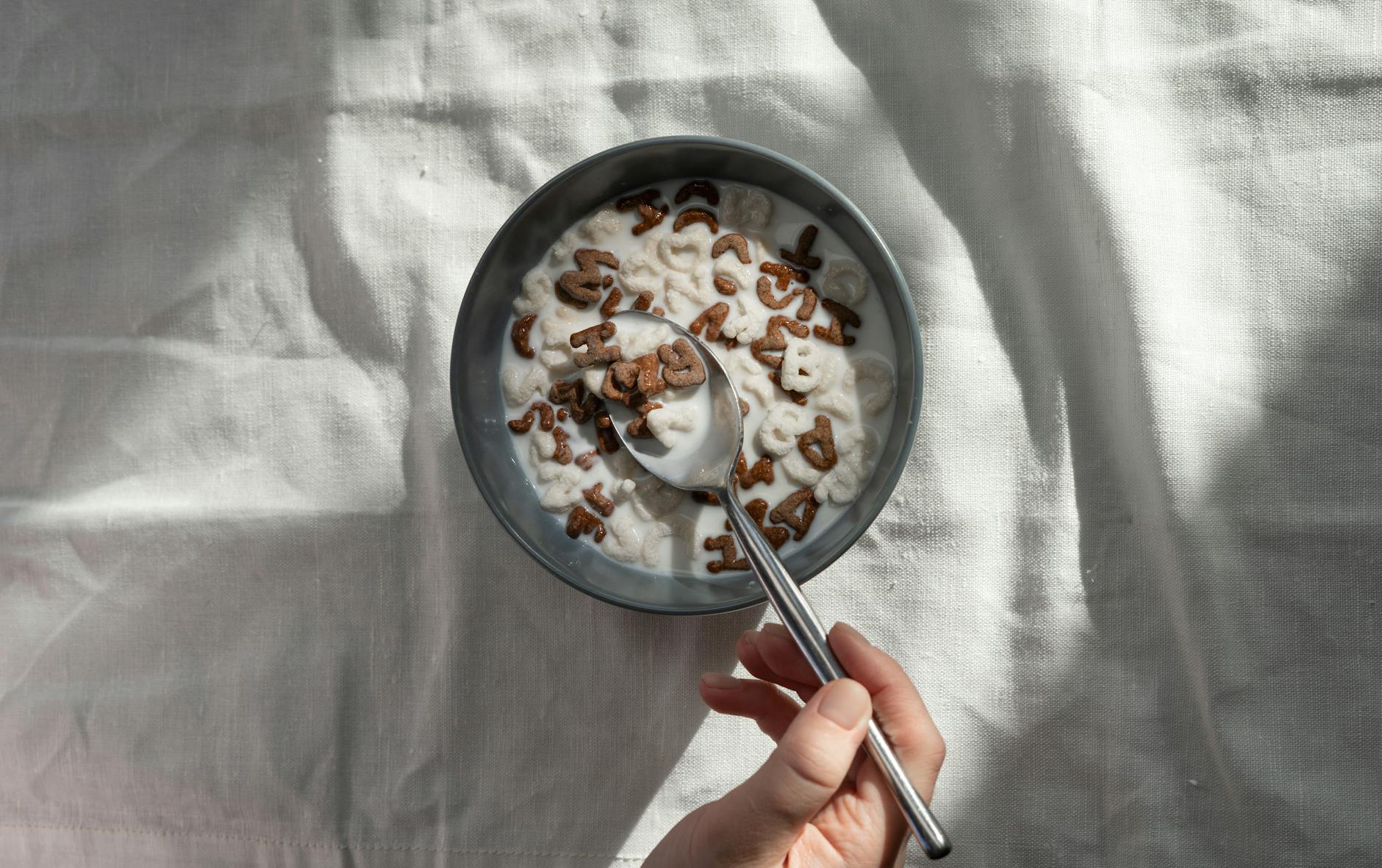 A close-up of a hand holding a spoon in a bowl of alphabet cereal and milk.