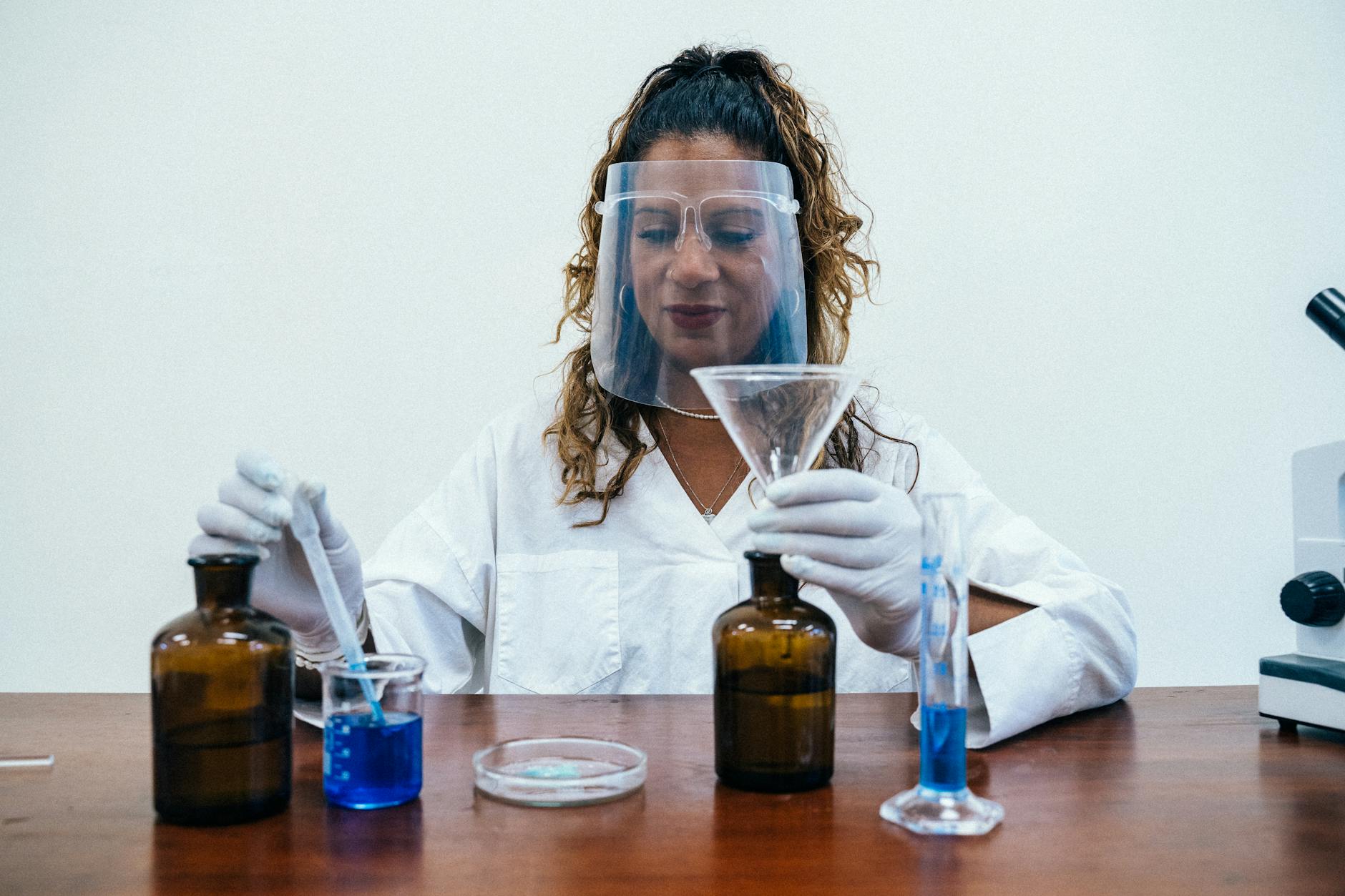 A female scientist in protective gear performs a lab experiment with blue liquid in containers.