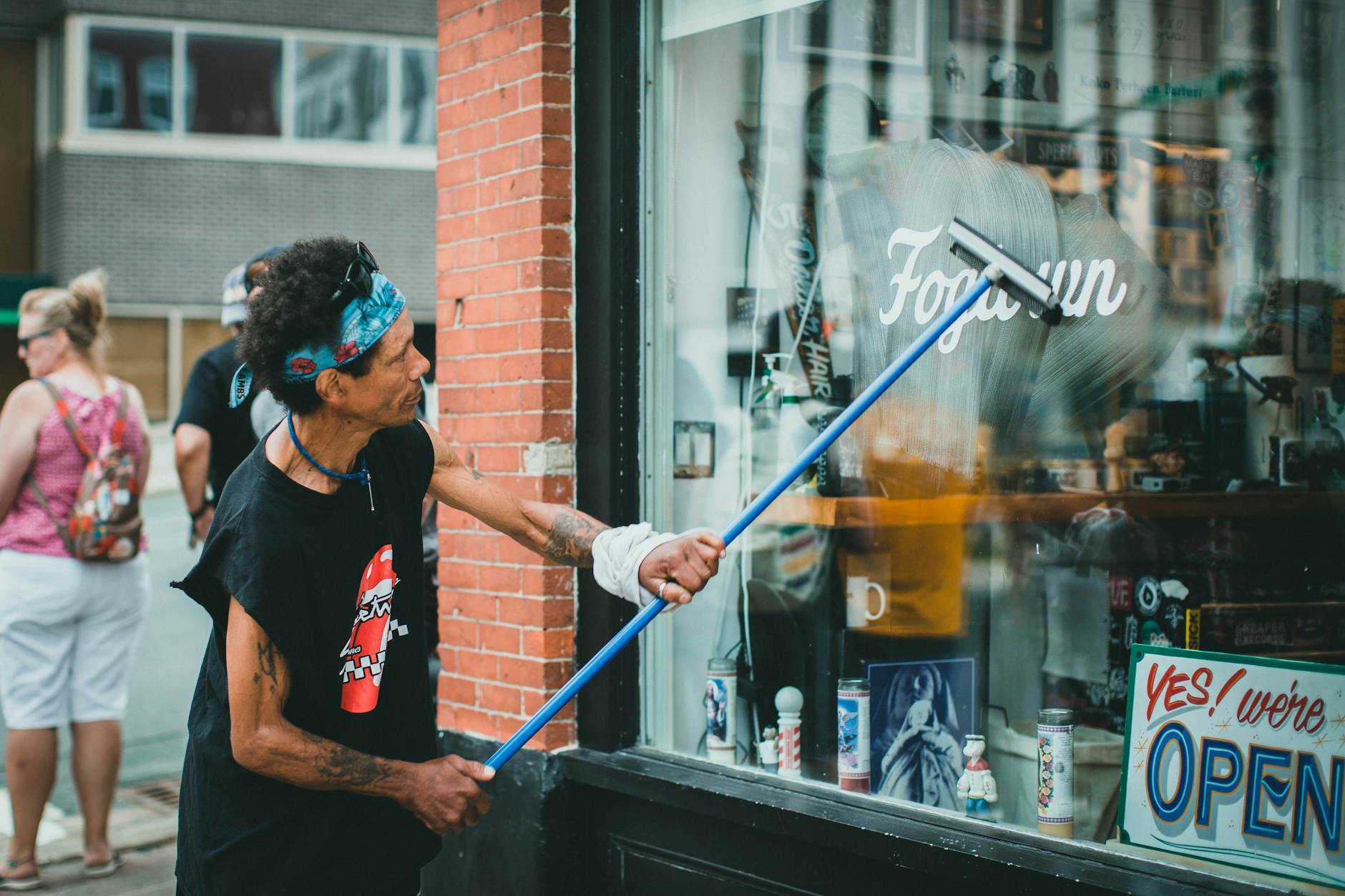 A man with tattoos cleaning a glass storefront window on a busy street using a squeegee.